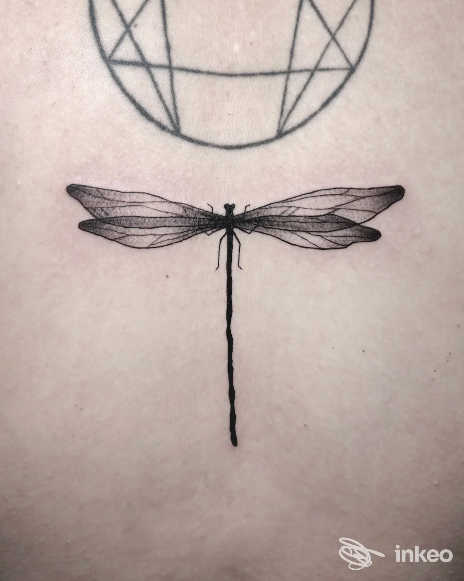 Tatuaggio realizzato sulla pelle raffigurante una libellula nera, con un simbolo geometrico circolare visibile sullo sfondo.
