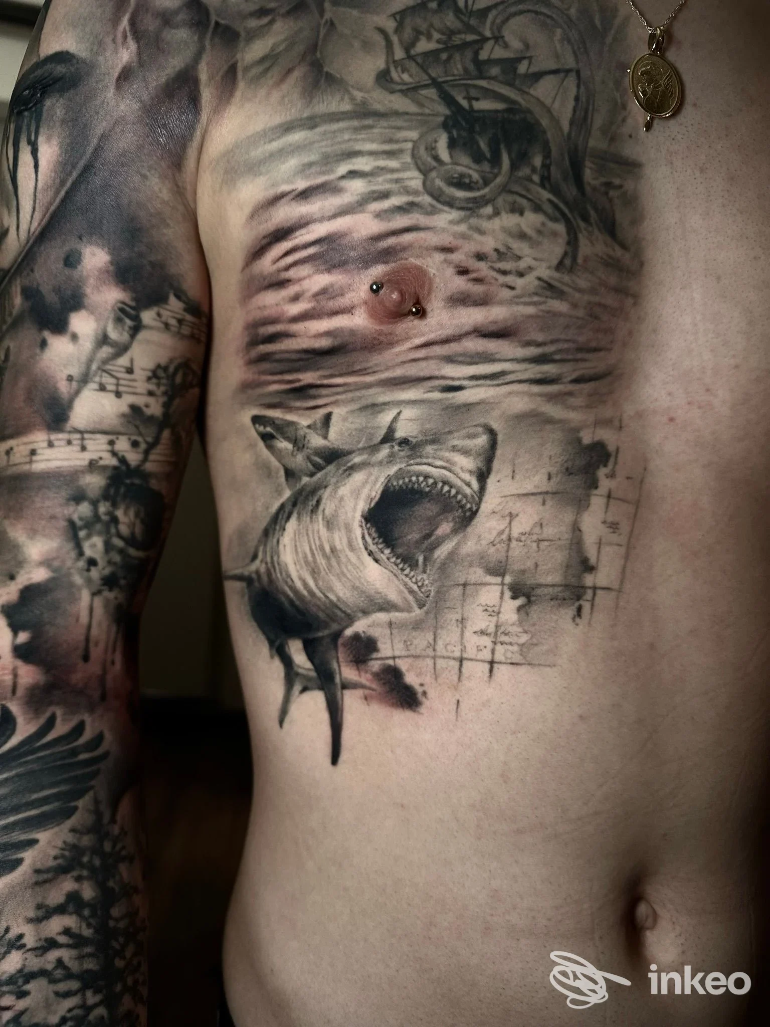 Tattoo realistica sul torace raffigurante uno squalo con dettagli in bianco e nero; piercing sul torace visibile, altri tatuaggi sul braccio sinistro. Ripresa singola.