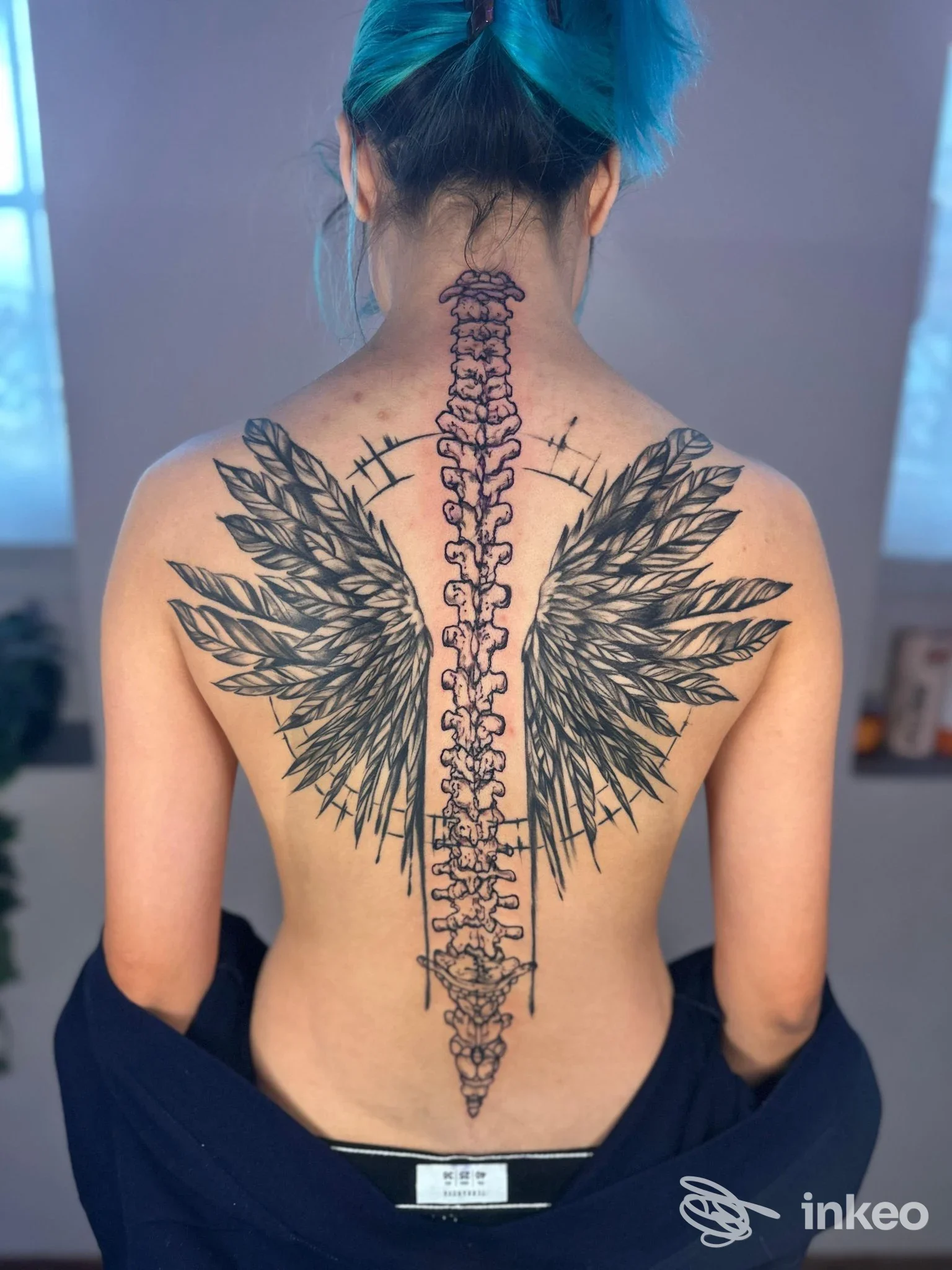 Schiena scoperta con grande tatuaggio centrale a colonna vertebrale e ali ai lati.