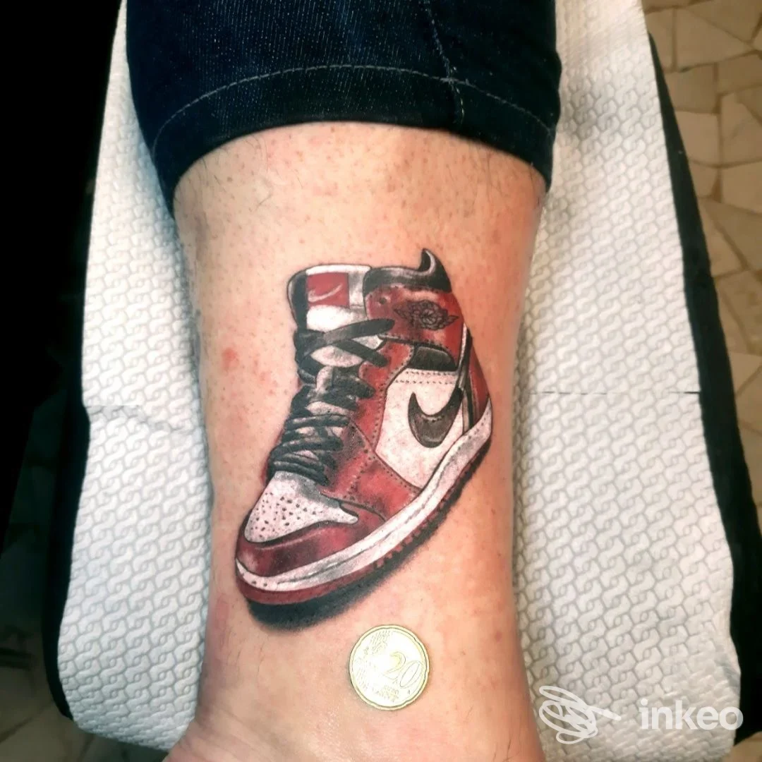 Tattoo realistico di una sneaker rossa e bianca sul polpaccio, con dettagli neri, realizzato su pelle umana; senza volto visibile o elementi d’interfaccia social.