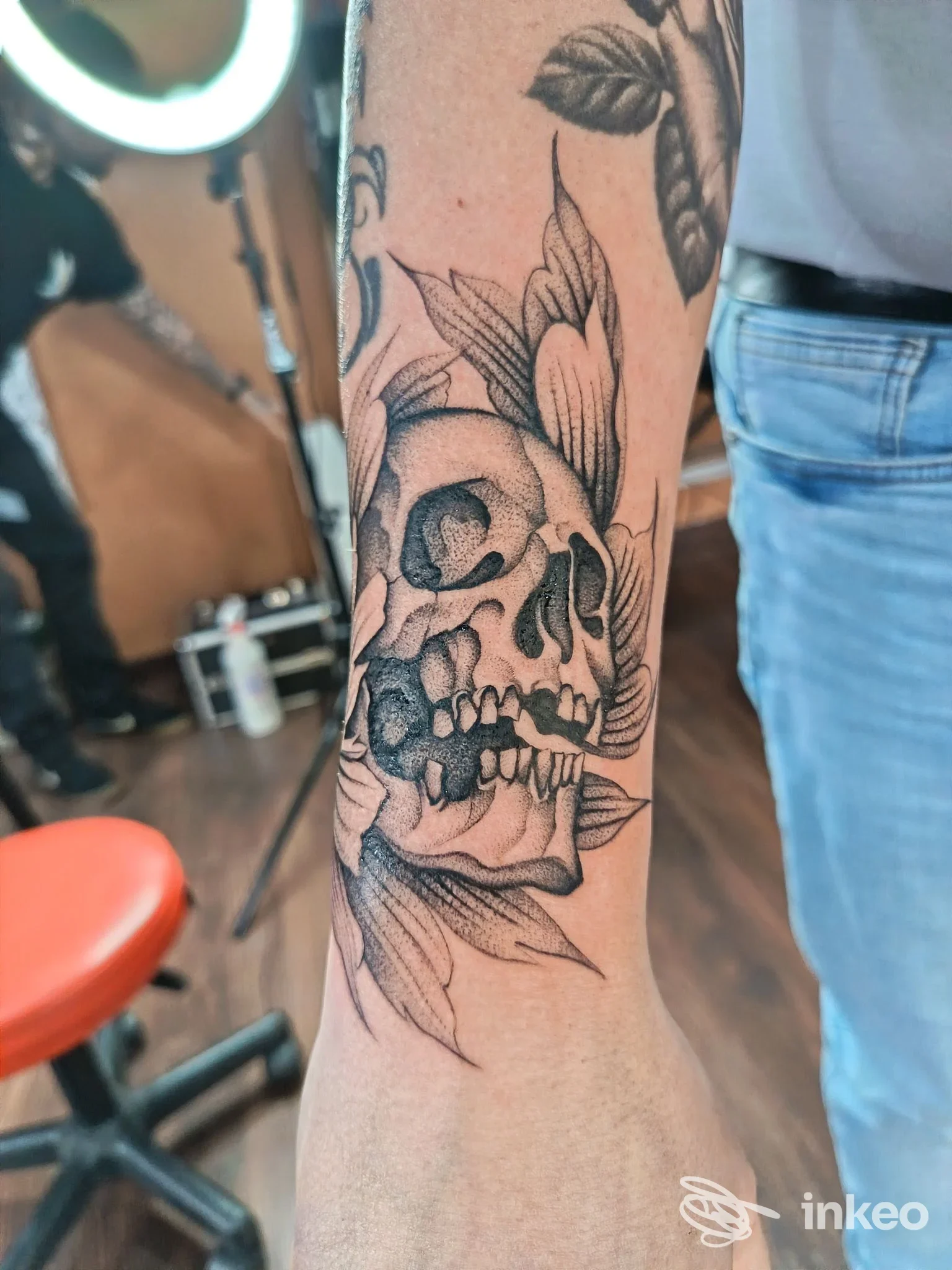 Tatuaggio su avambraccio destro: un teschio circondato da foglie e fiori in tonalità nere/grigie, realizzato con linee nette e shading.