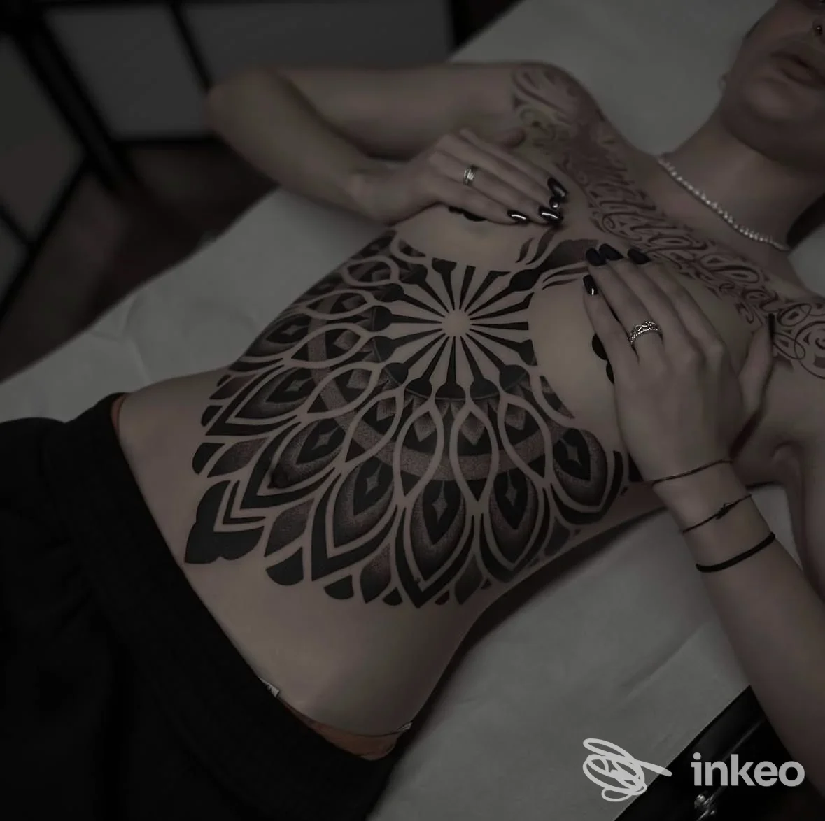 Tattoo geometrico-ornamentale nero copre torace e addome di una persona sdraiata, con mani visibili che ostentano anelli; immagine promossa da studio Manomorta Tattoo.