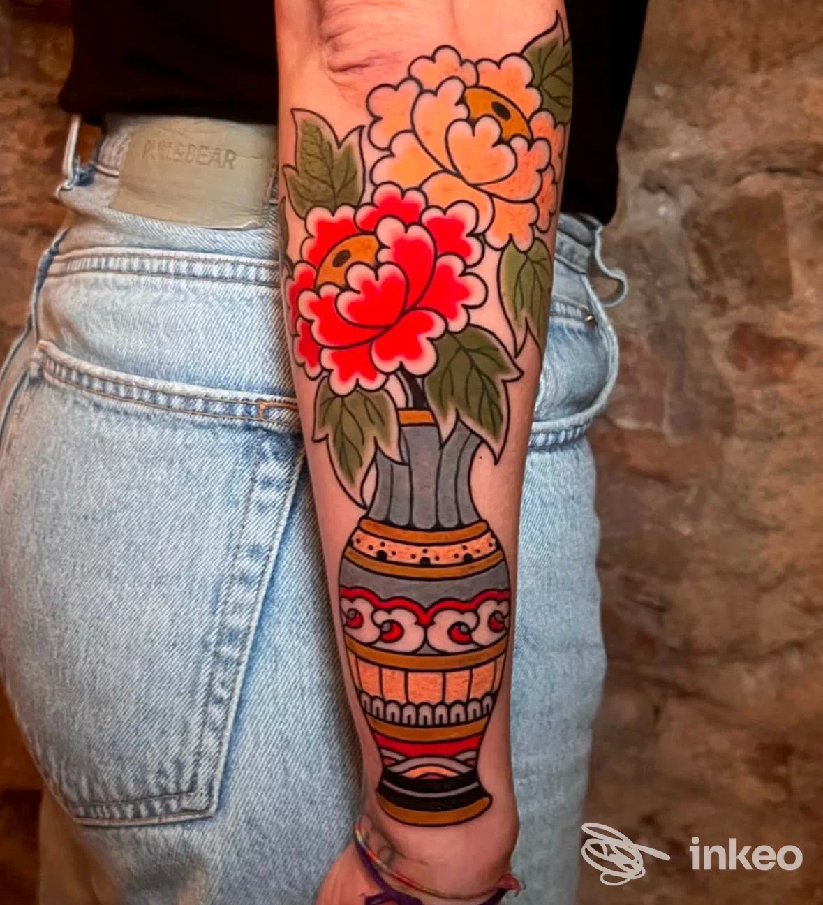 Tattoo a penelope sull'avambraccio raffigurante un vaso decorato con grandi gigli rossi e rosa, con foglie verdi e motivi geometrici, sfondo parete di pietra, scatto frontale. Studio Manomorta Tattoo.