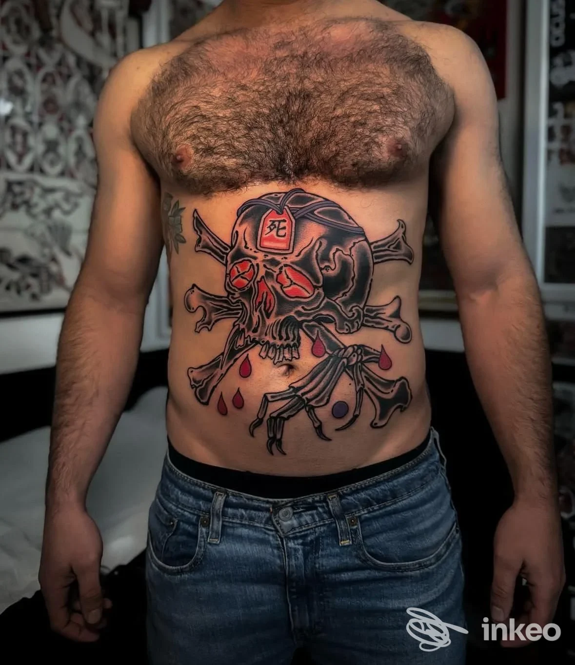 Soggetto maschile con grande tatuaggio sull’addome raffigurante un teschio con ossa incrociate e gocce di sangue; torace nudo, jeans visibili in vita.