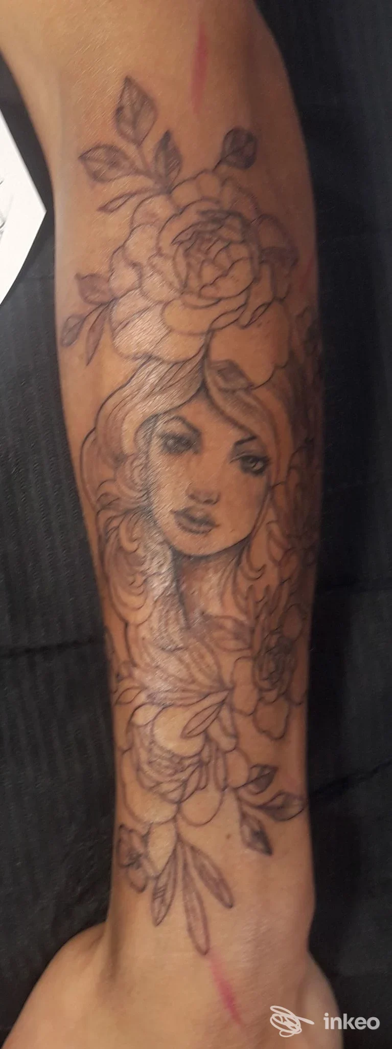 Tatuaggio visibile sull'avambraccio raffigurante una donna circondata da bouquet di fiori, tratteggiato in bianco e nero su pelle. studio One Way Tattoo a Usmate Velate.