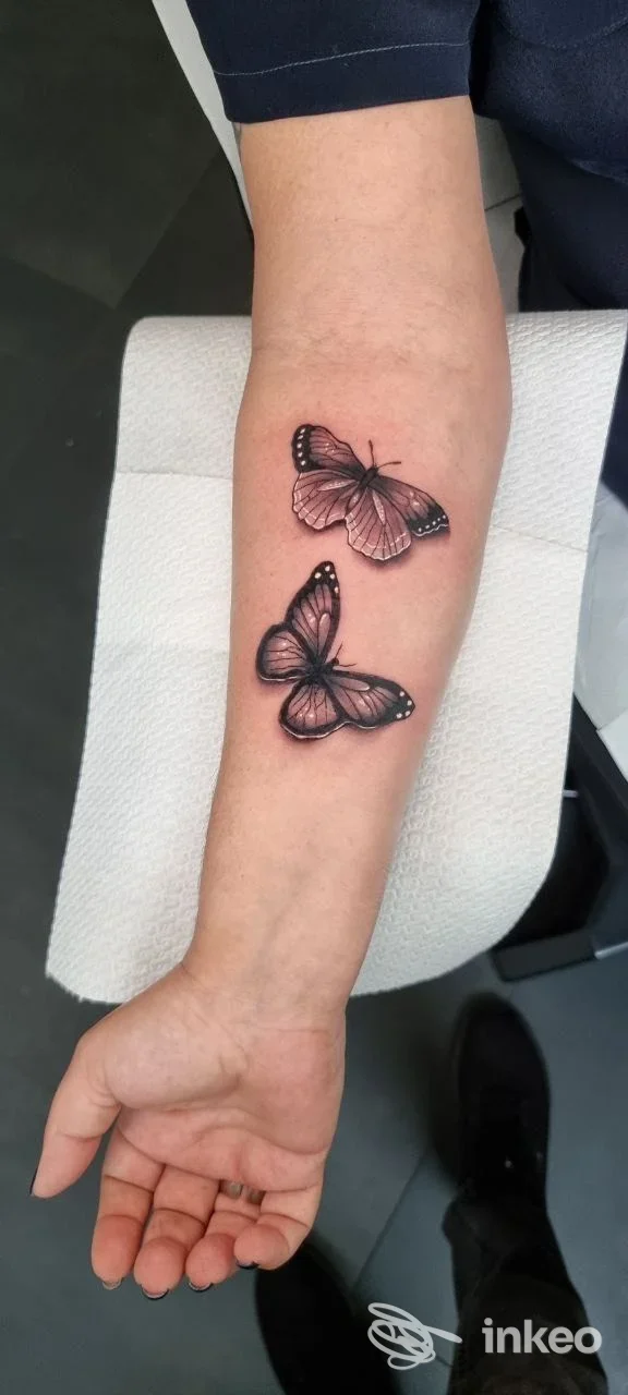 Avambraccio sinistro con due farfalle tattoo, pelle reale ben visibile, colori rosa e neri, nessun volto in scena, nessuna sovraimpressione.