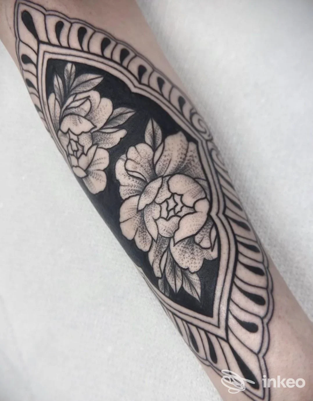Braccio con tatuaggio floreale su sfondo scuro, contorni geometrici e cornice, disegno in bianco su pelle chiara, stile ornato.