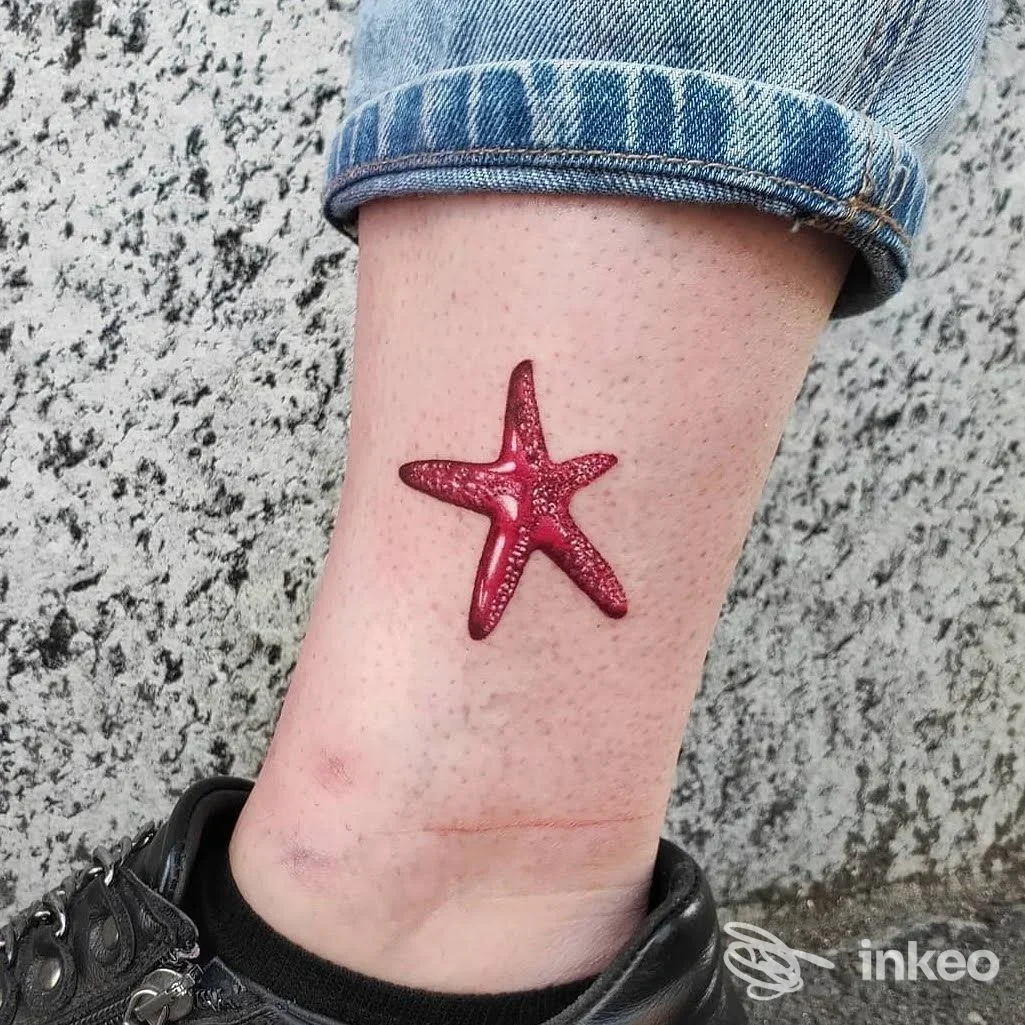 Tatuaggio a stella marina rosso glitterato sulla caviglia di una persona, con jeans e scarpa scura visibili.