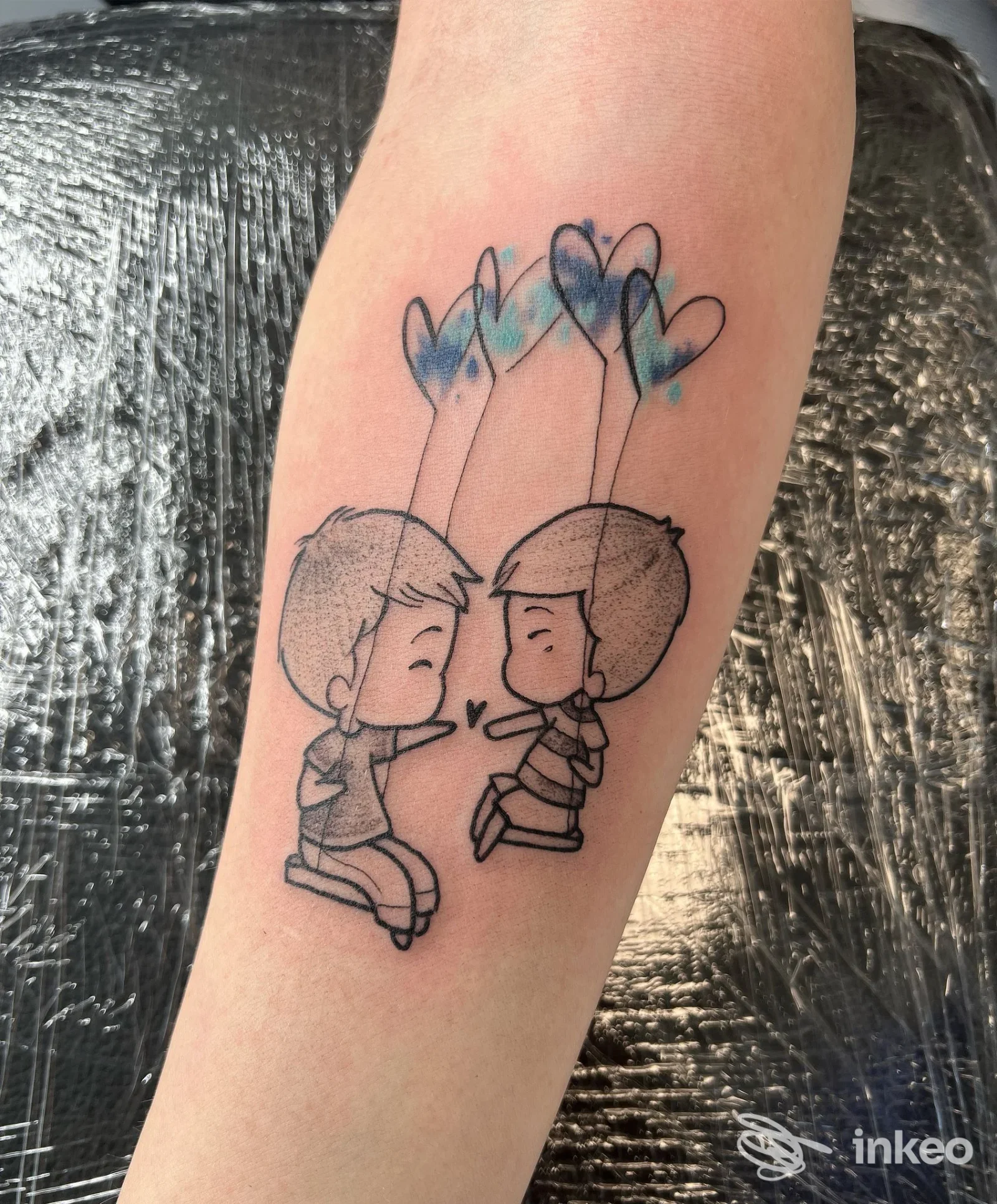 Tatuaggio su avambraccio raffigurante due bambini in stile cartone animato che si sfiorano con cuori azzurri sopra, realizzato sulla pelle senza presenza di volto umano visibile.