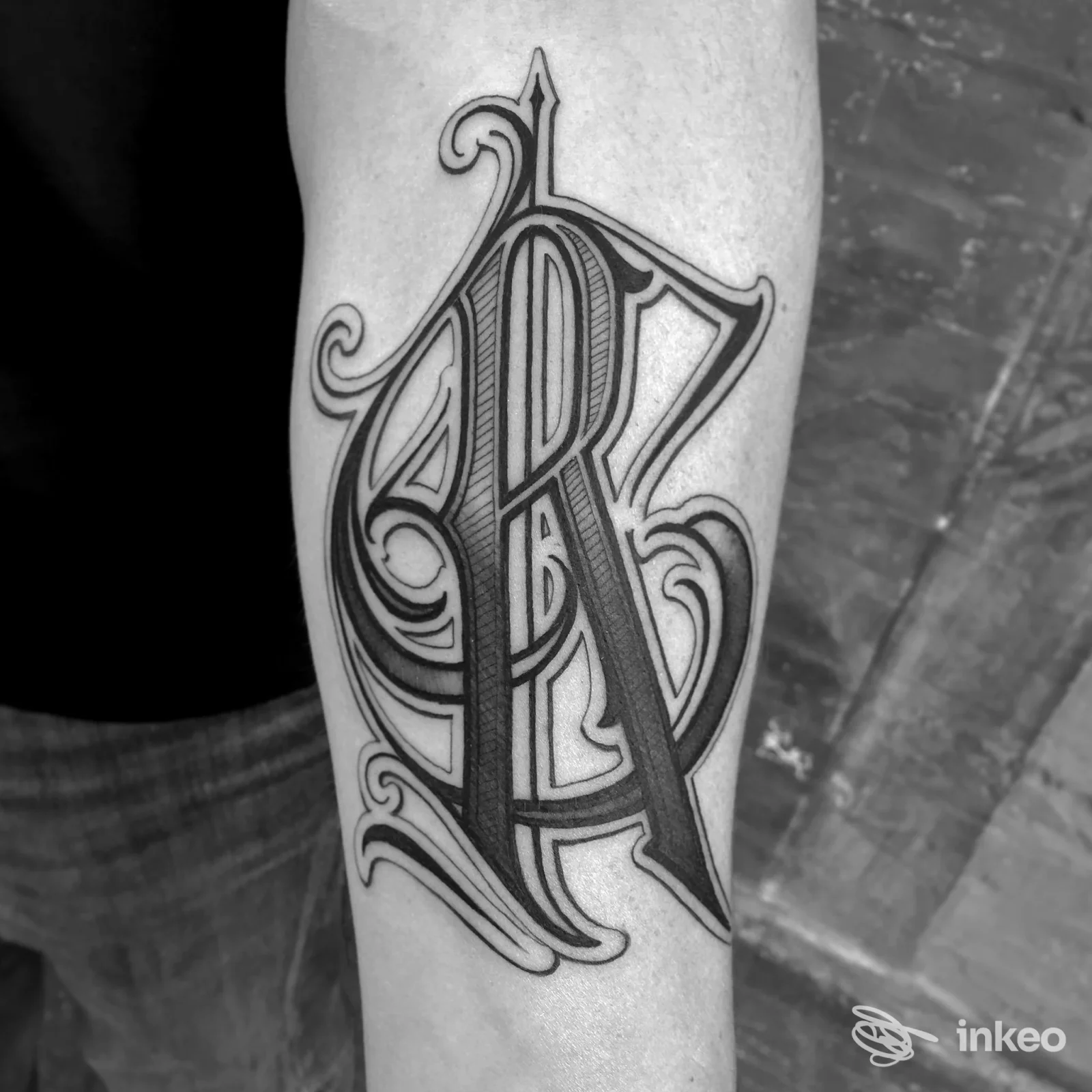 Tatuaggio ornato in stile lettering su avambraccio, linee scure con riempimenti sottili su pelle chiara, scatto frontale della zona tatoo.