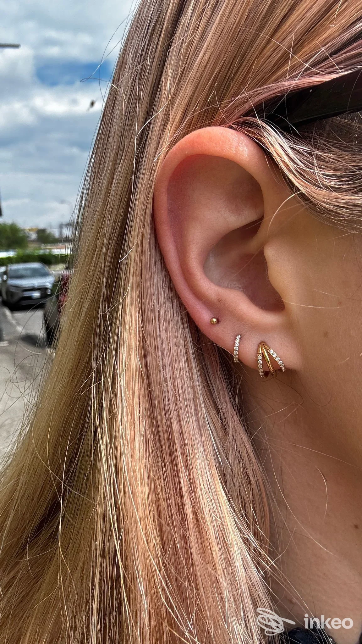 Primo piano di un orecchio con due piercing: un piccolo stud sul lobulo e un orecchino decorato in doratura, con capelli chiari in background.