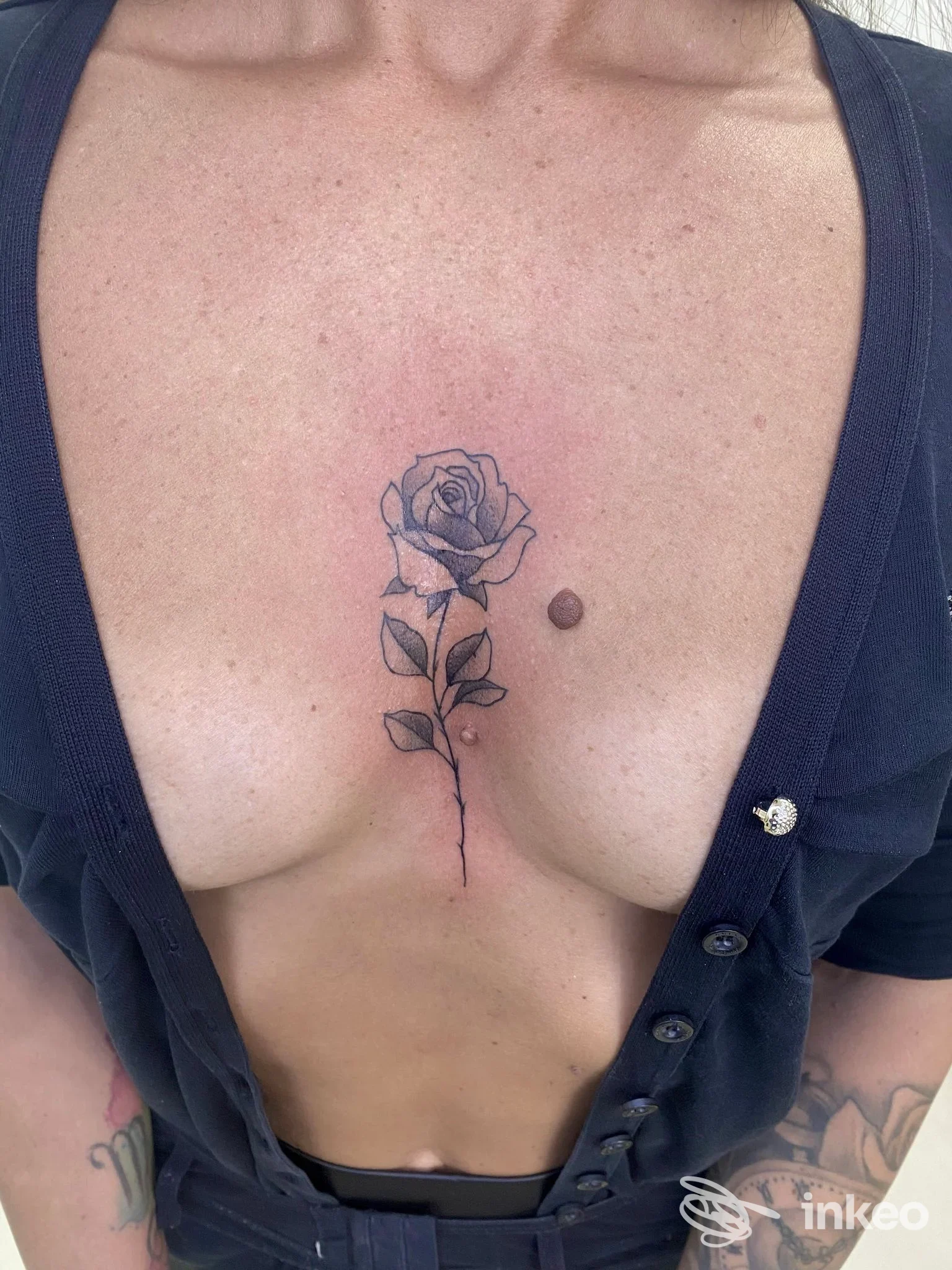 Tattoo visibile di una rosa lungo lo sterno, disegno su pelle umana, visibile dal décolleté, con tonalità di blu e verde. Studio SG Studio, località non specificata.