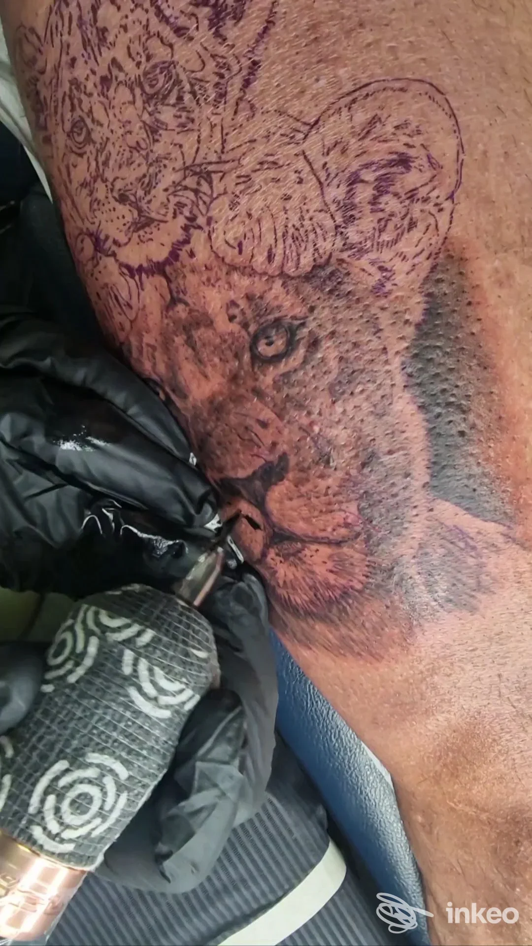 Immagine di una sessione di tatuaggio: una macchina per tatuare è in uso su pelle con disegno viola in corso; non si vede il volto del cliente.