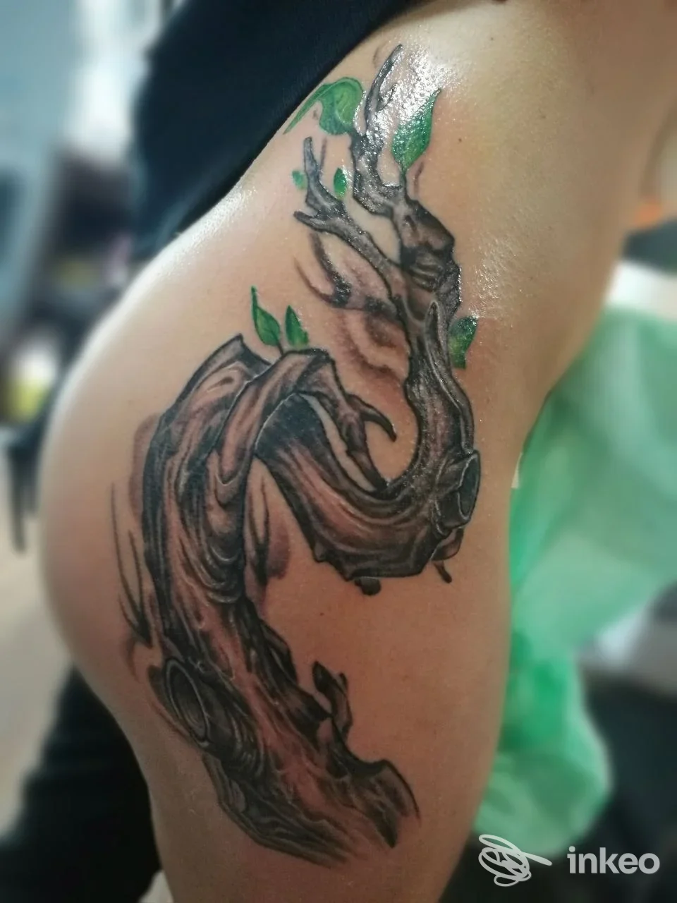Tatuaggio su braccio raffigurante un ramo contorto in tonalità scure con qualche foglia verde, stile realistico.