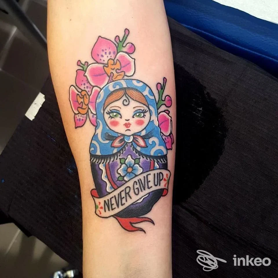 Tatuaggio colorato sull'avambraccio raffigurante una bambolina in stile Matryoshka con fiori e un nastro con la scritta Never Give Up, realizzato sulla pelle.