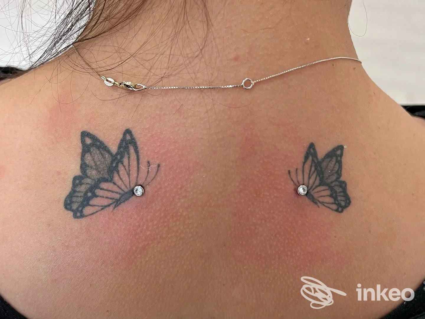 Due tatuaggi a farfalla sulla nuca, ognuno con una piccola gemma brillante; la pelle circostante è leggermente arrossata.