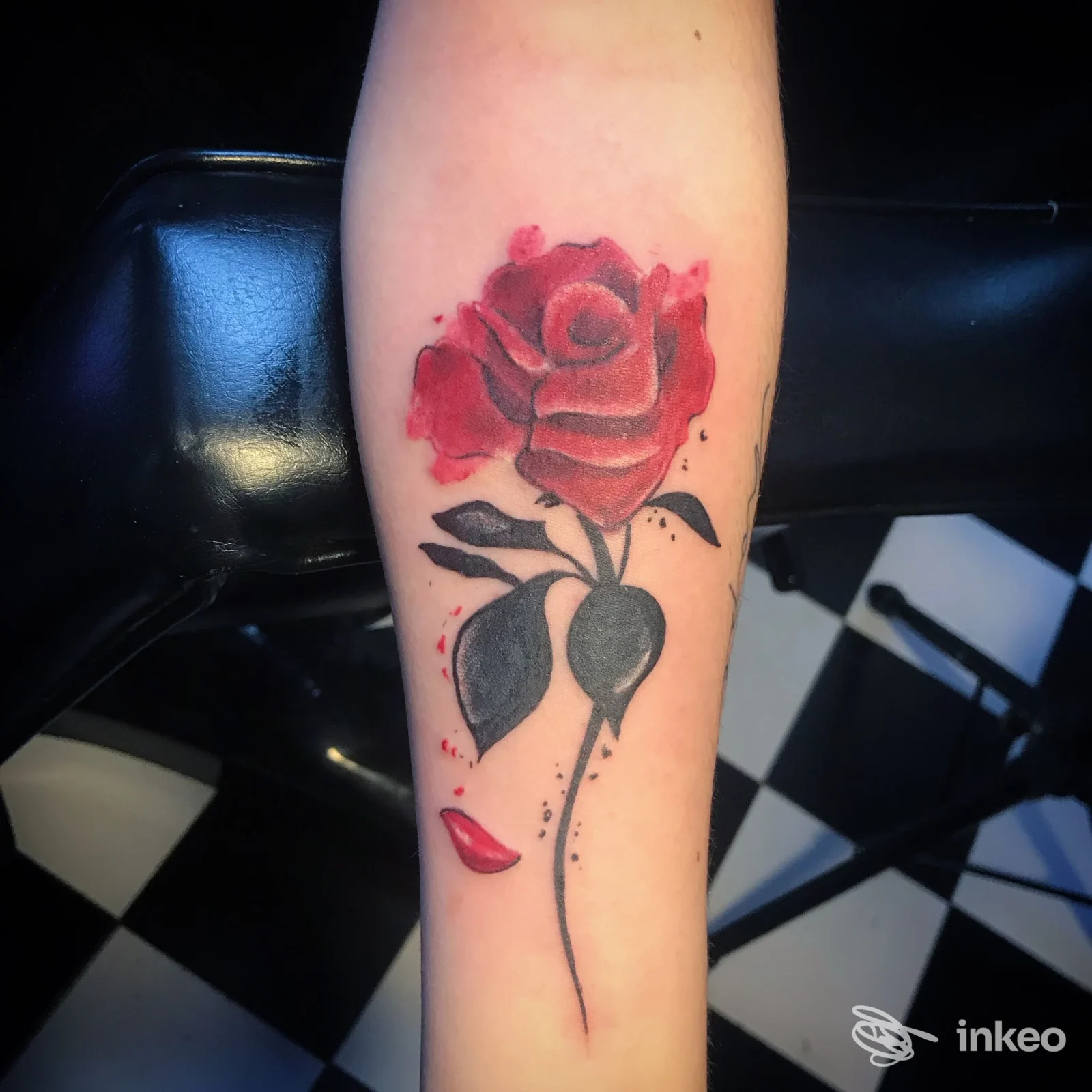 Tattoo su avambraccio raffigurante una rosa rossa con foglie, eseguita sulla pelle, sfondo con pavimento a scacchi.
