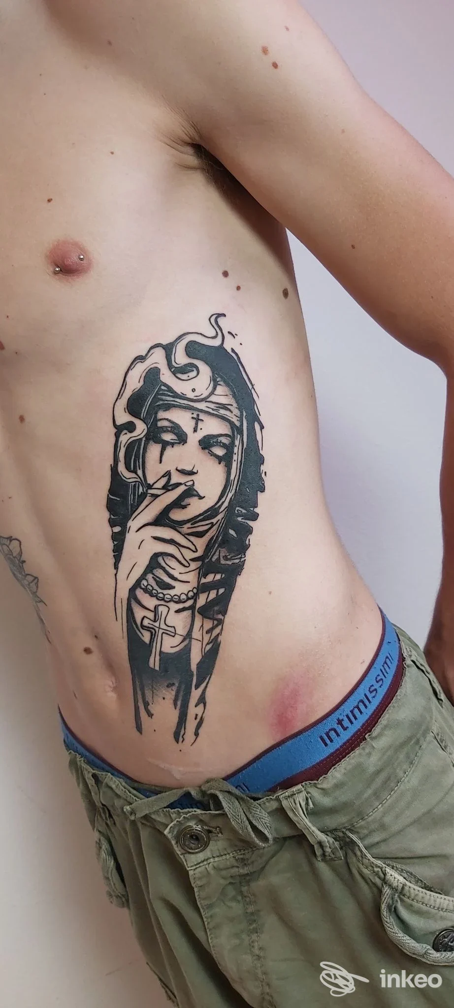 Tattoo verticale su fianco destro di una figura femminile con mantello, visibile parte del torace; la persona è in biancheria e pantaloni verdi, nessun testo sovrapposto.