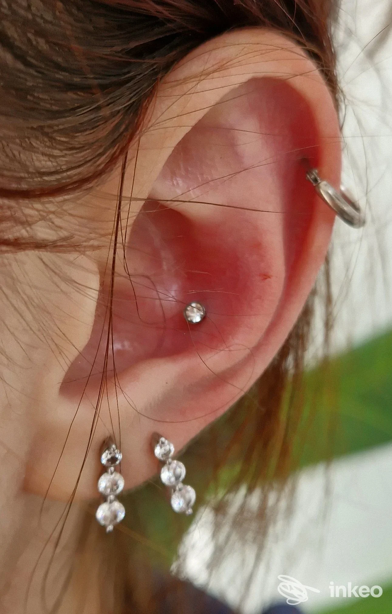 Orecchio sinistro con piercing: helix superiore con cerchietto metallico e stud al lobo, accompagnato da orecchini pendenti. Parte di capelli visibile.