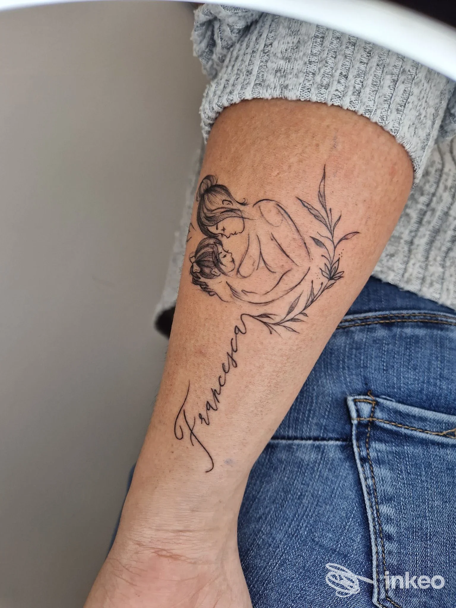 Braccio sinistro di una persona con tatuaggio visibile: una mano stilizzata trattiene una foglia o fiore, accompagnata da una scritta corsiva lungo l’avambraccio; nessun volto visibile.