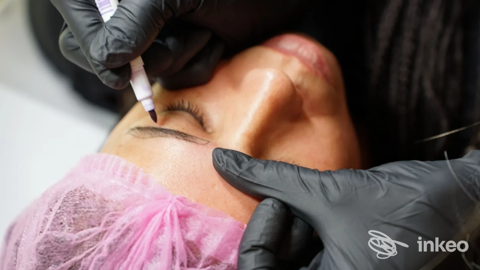 Procedura di trucco permanente sull’arcata sopraccigliare/occhio: mani guantate e una penna per tatuaggio vicino alla pelle della persona, con cuffia rosa in primo piano.