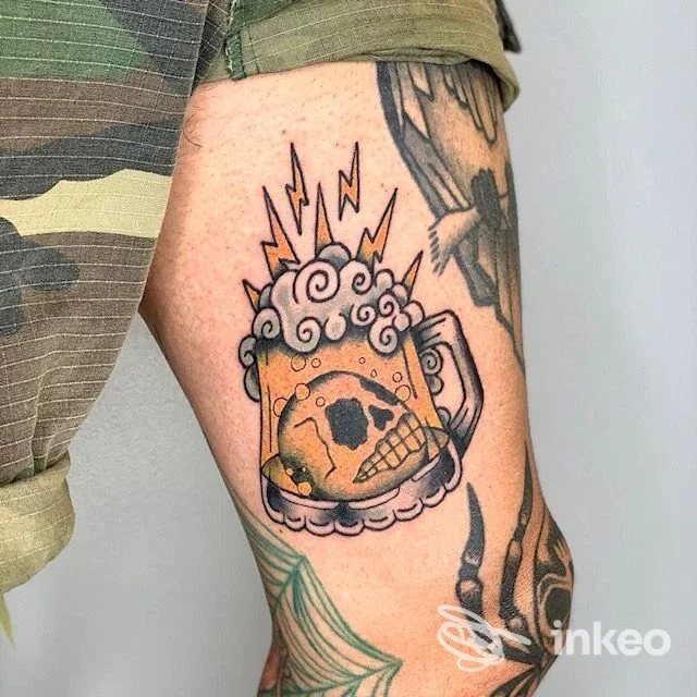 Tattoo sull'avambraccio raffigurante una tazza di birra con teschio, contorni neri e colori arancione-giallo, parte di un braccio coperto da tatuaggi.
