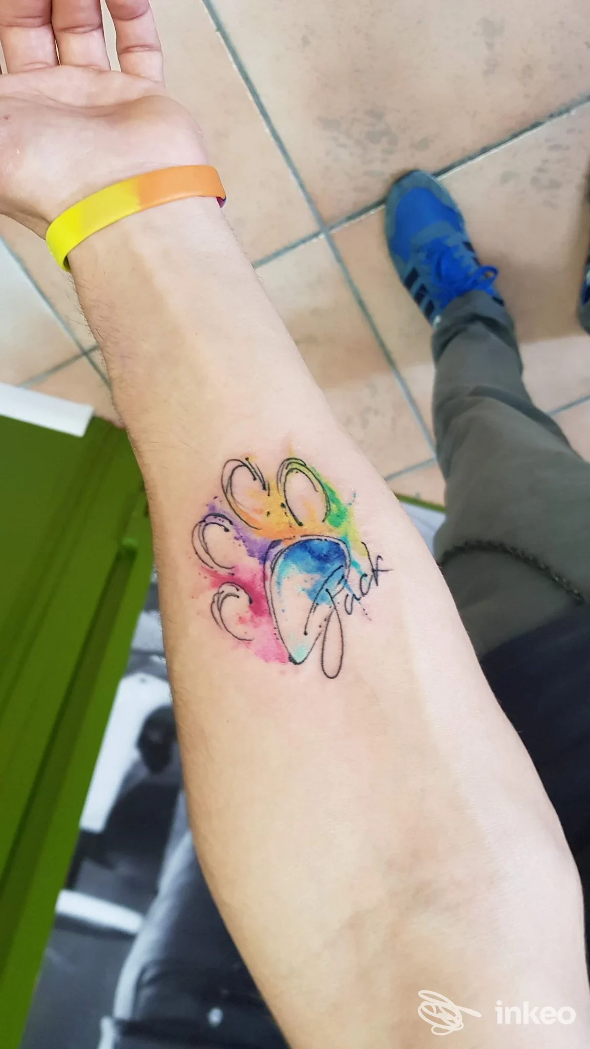 Braccio con tatuaggio colorato ad acquerello a cuore e motivi floreali, visibile sulla pelle; fondo pavimentato con persona in pantaloni grigi e scarpe blu.