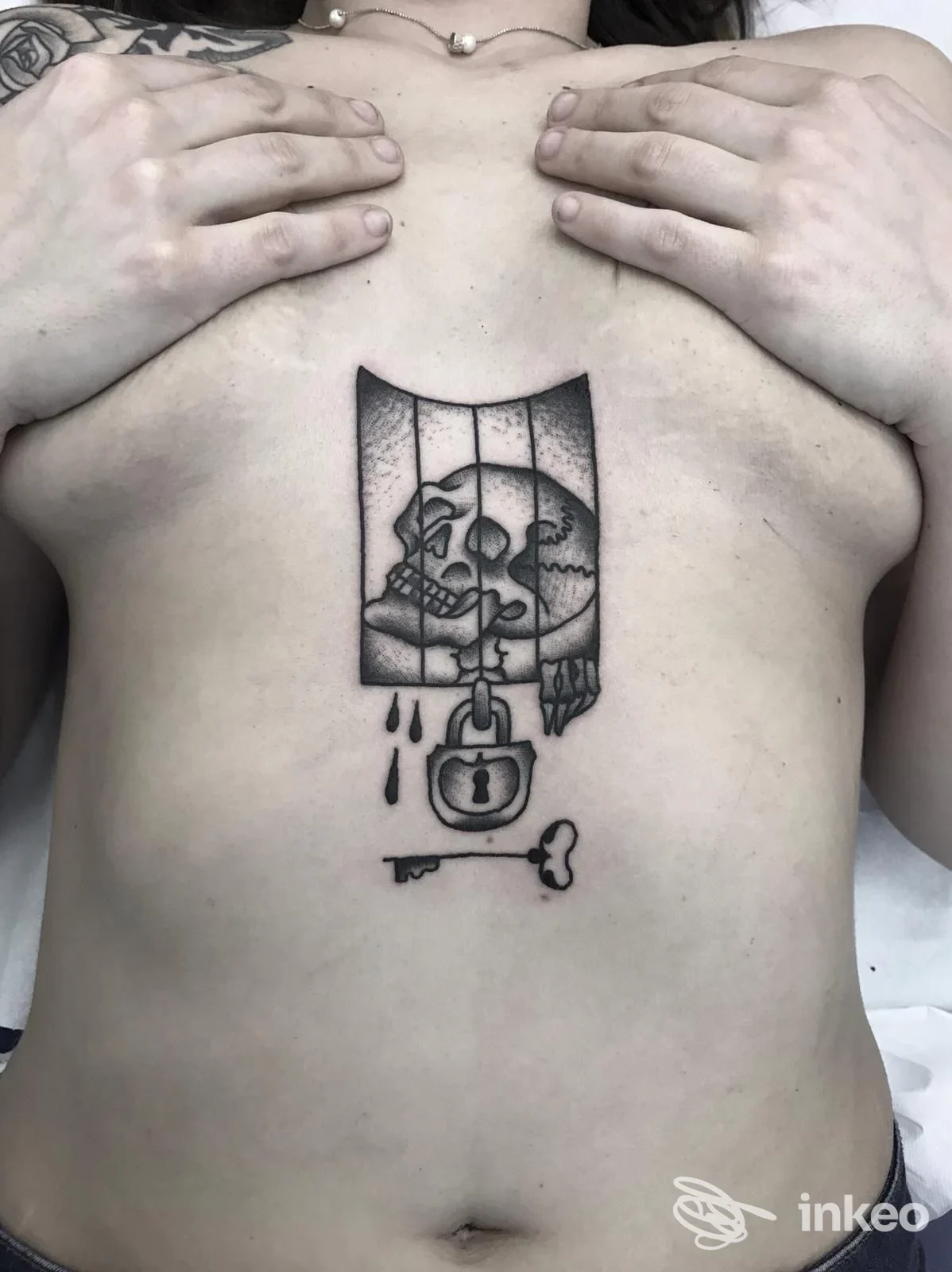 Tattoo sul torace raffigurante un teschio in una gabbia con lucchetto e chiave, mani inginose ai lati, foto singola.
