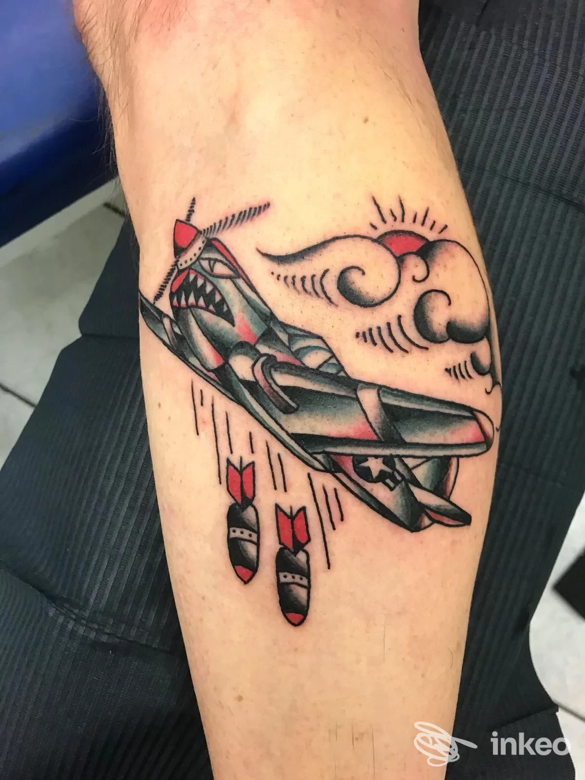 Tatuaggio su avambraccio raffigurante un aereo da guerra in stile tradizionale, con bombe che cadono e nuvole sullo sfondo, realizzato sulla pelle.