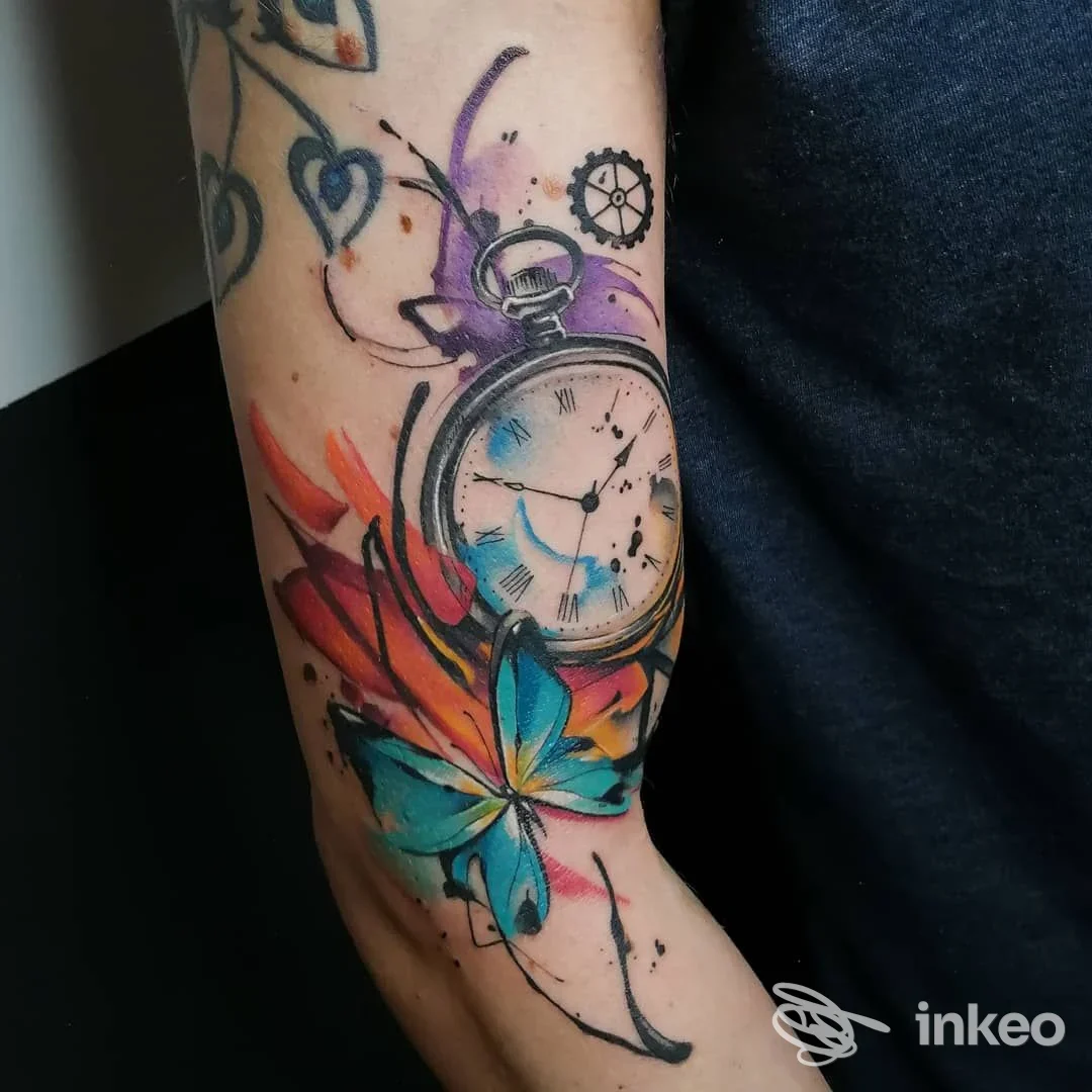 Tatuaggio a braccio raffigurante un orologio a pendolo con colori vivaci e una farfalla, su pelle reale. Studio Youcanvas Tattoo Studio, immagine non promozionale.