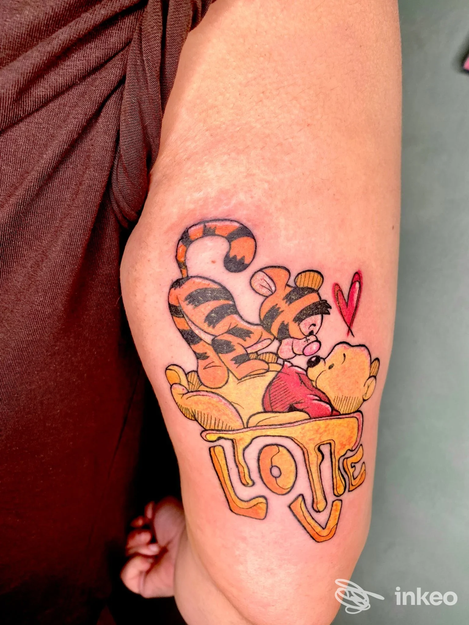 Tattoo raffigurante Winnie the Pooh e Tigger su avambraccio, colori vividi e linee pulite, con cuore rosso vicino e uno sfondo di pelle naturale.