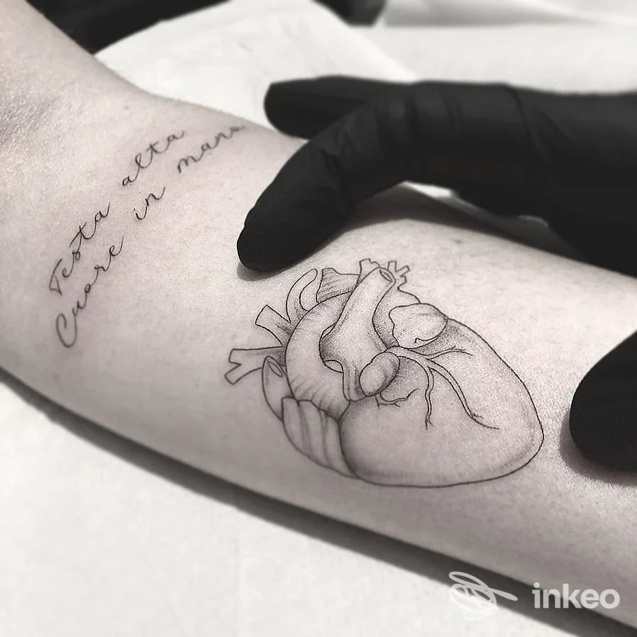 Diplomando tatuaggio reale sull'avambraccio raffigurante un cuore anatomico in linea nera, con una mano guantata che lo sfiora. Si intravede anche una scritta a lato.