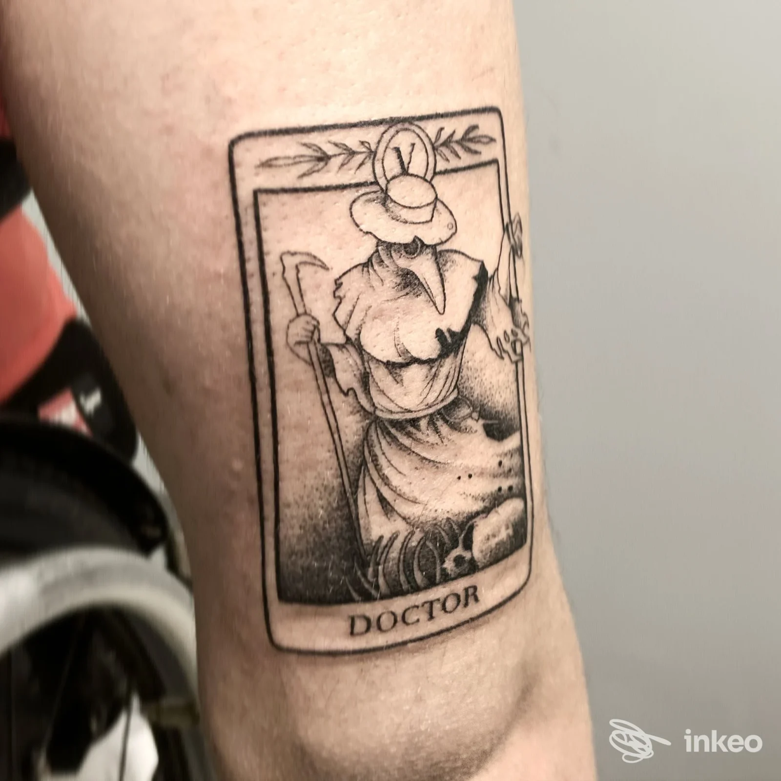 Tatuaggio su avambraccio raffigurante una figura in stile medico della peste all’interno di una cornice, con la parola DOCTOR in fondo, eseguito su pelle.